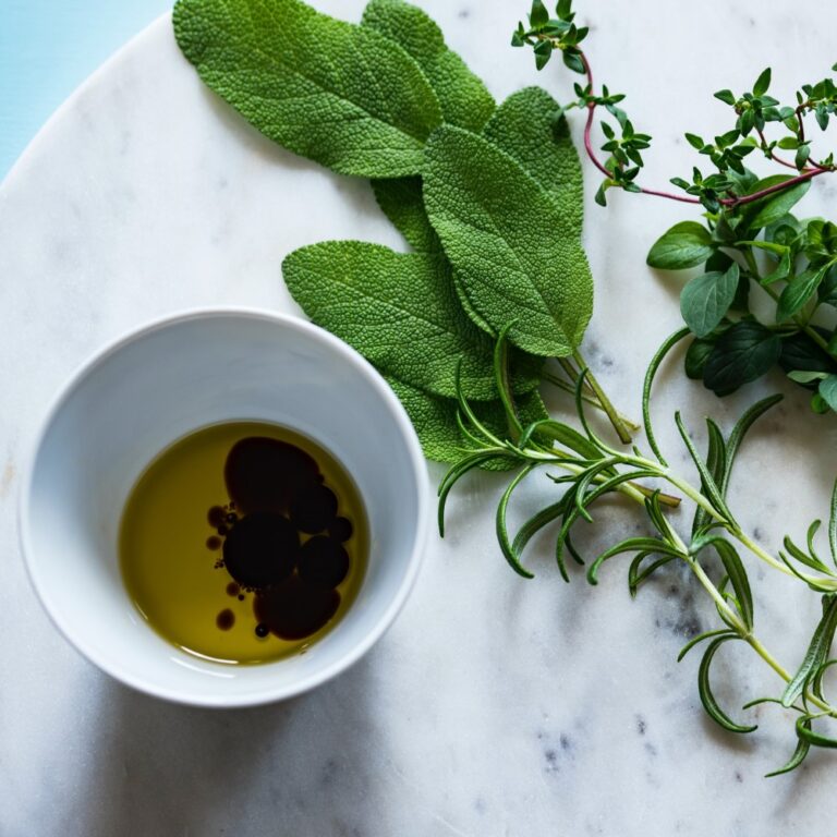 overhead view of white bowl with oil, balsamic vinegar and fresh herbs on a marble tabletop